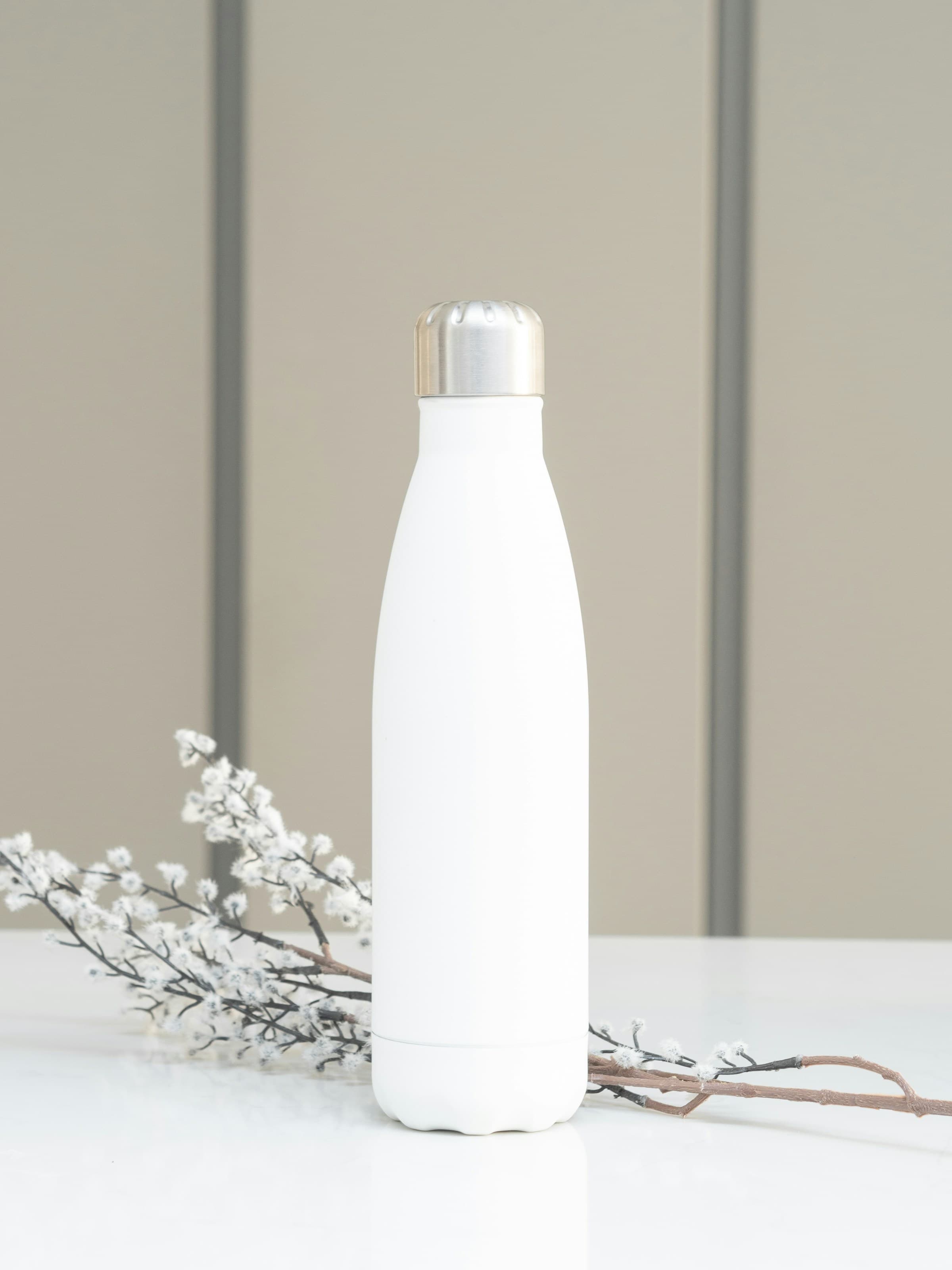 A matte white insulated bottle with a brushed steel cap, standing beside a sprig of dried blossom.