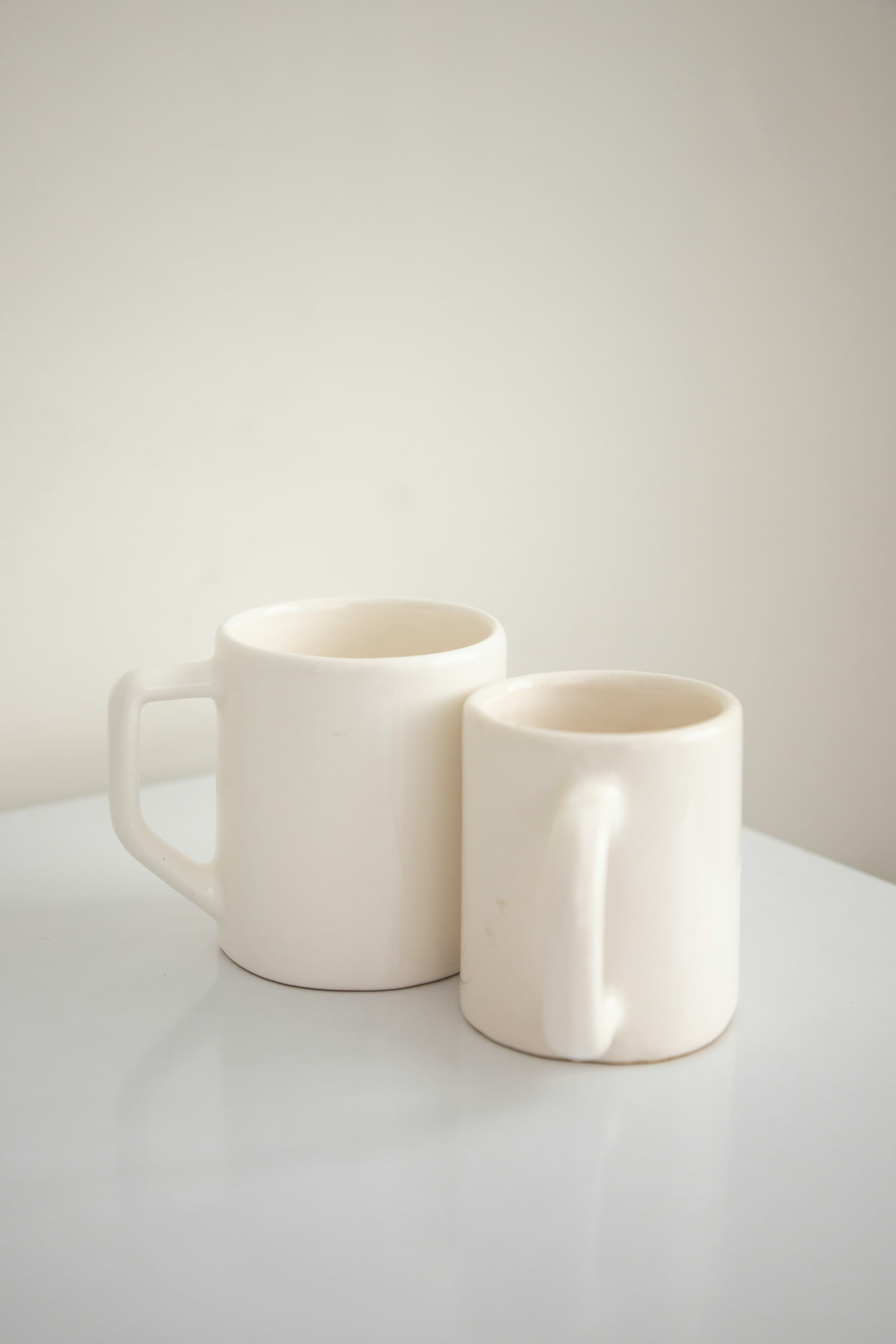 Two off-white ceramic mugs of different heights on a pale table.