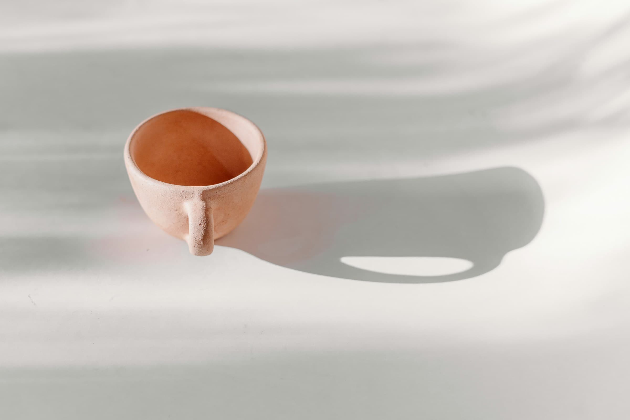 A small terracotta cup casting a long shadow across a white surface in soft daylight.